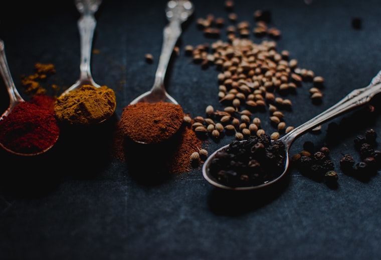 Spoons of ground spices and whole peppercorns on dark slate